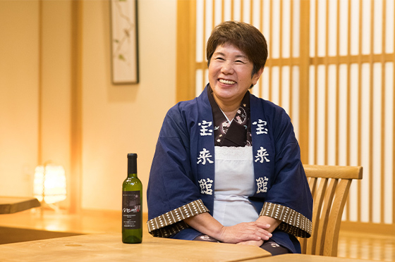 老舗旅館・宝来館のロビーにて、オリジナルワインのマロードを手元に笑顔でワインプロジェクトのことを語る岩崎昭子さん