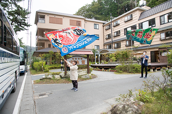 朝の宝来館の前に2台の観光バスが並ぶ。中には30名近いクボタスピアーズのラグビー選手らが乗車しており、宝来館の女将・岩崎さんが彼らに向かって釜石ラグビーの名物・大漁旗を力いっぱい振ってエールを送っている