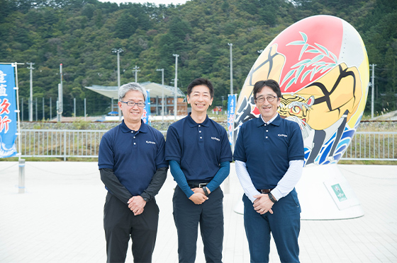 釜石市を縦断する三陸鉄道の駅のひとつ鵜住居駅の前の広場にて、クボタの服部竜一さん、菱田真さん、堀越健文さんが笑顔で記念写真におさまる