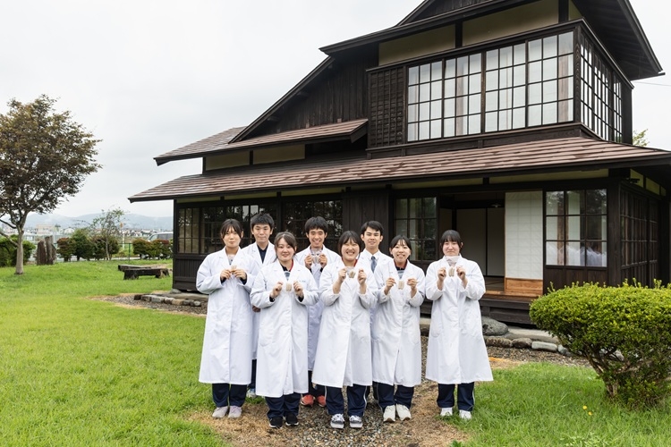 高校の敷地内にある宮沢賢治ゆかりの建物「羅須地人（らすちじん）協会」の前に集まり、商品化されたソーセージを掲げるソーセージ研究班の生徒たち