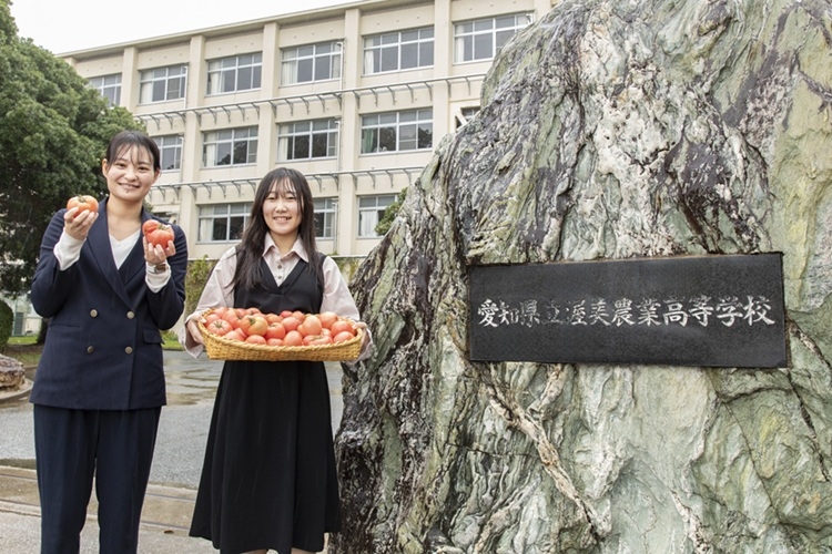 渥美農業高等学校の正門前でトマトを手に持つ元動物科学部の卒業生と元顧問の先生