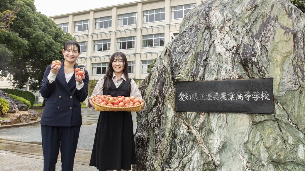 渥美農業高等学校の正門前でトマトを手に持つ元動物科学部の卒業生と元顧問の先生
